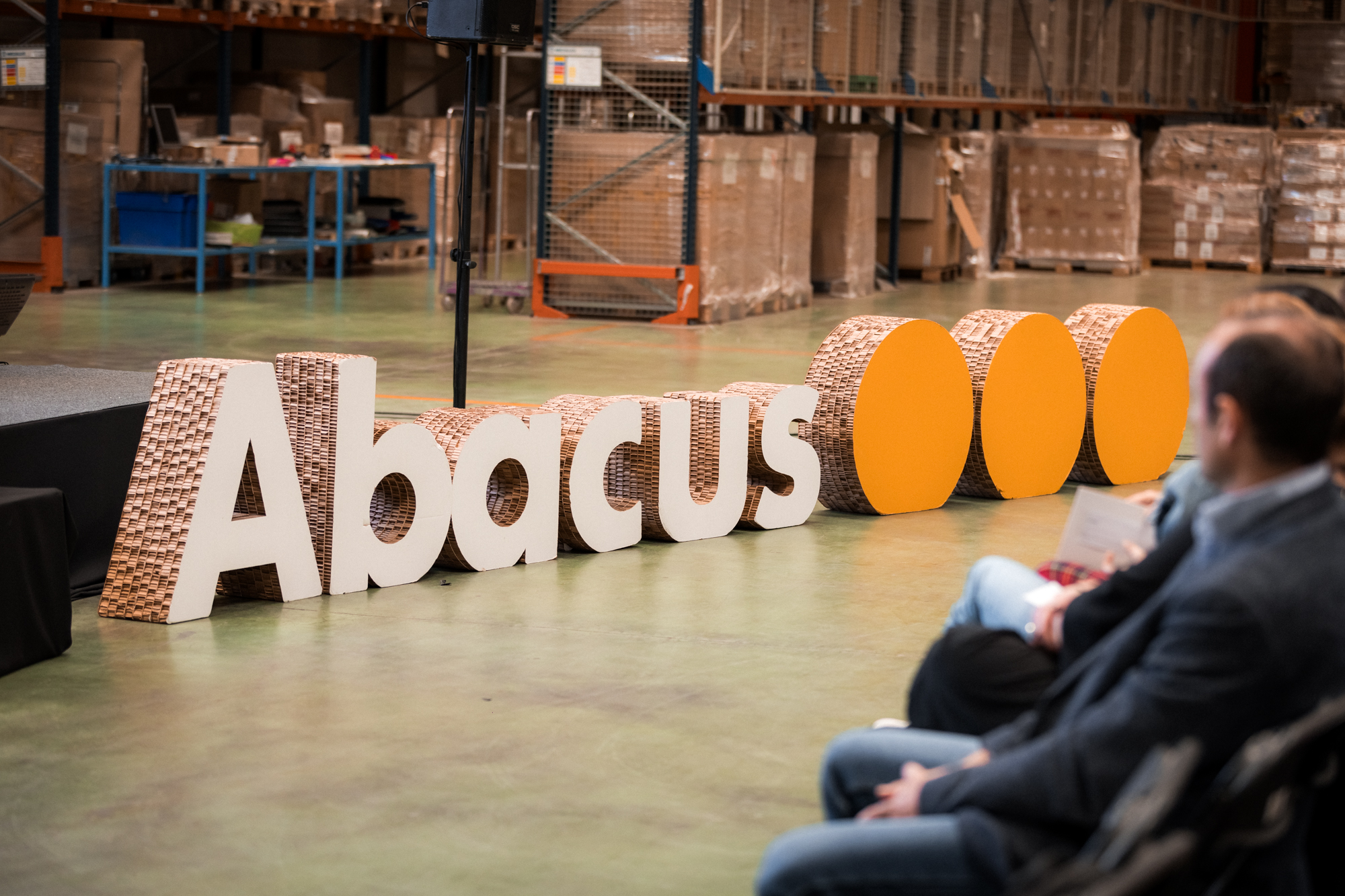 Imagen de una de las asambleas realizadas en el centro logístico de abacus, se ve parte del escenario y el logotipo de abacus realizado con piezas de cartón.